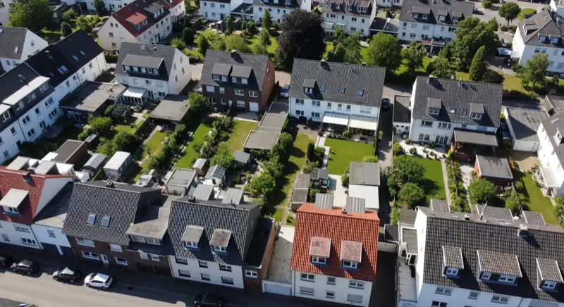 Aerial drone view of typical German residential neighborhood with mixed roof types, red and dark roof tiles, gardens visible, sunny day