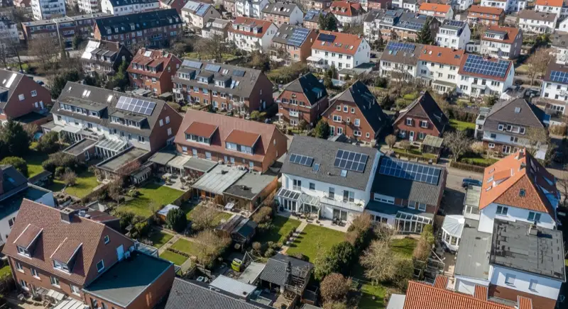 Aerial drone view of typical German residential neighborhood with mixed roof types, red and dark roof tiles, gardens visible, sunny day
