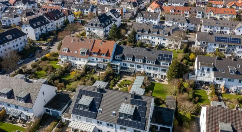 Aerial drone view of typical German residential neighborhood with mixed roof types, red and dark roof tiles, gardens visible, sunny day