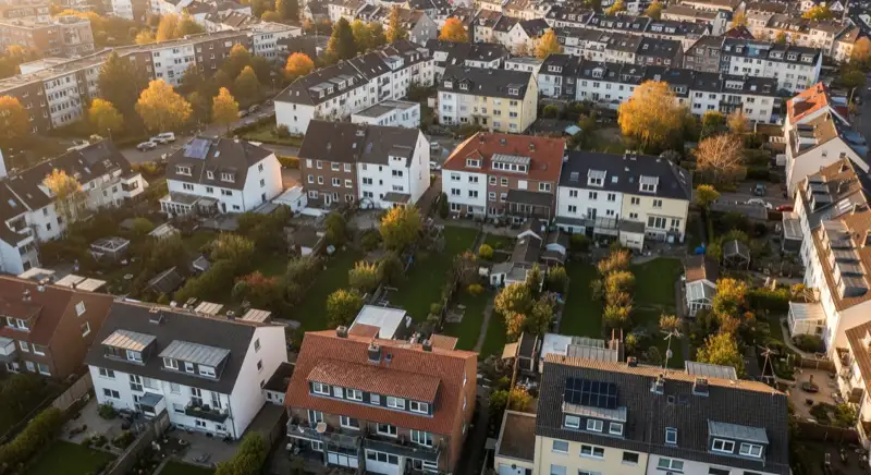 Aerial drone view of typical German residential neighborhood with mixed roof types, red and dark roof tiles, gardens visible, sunny day