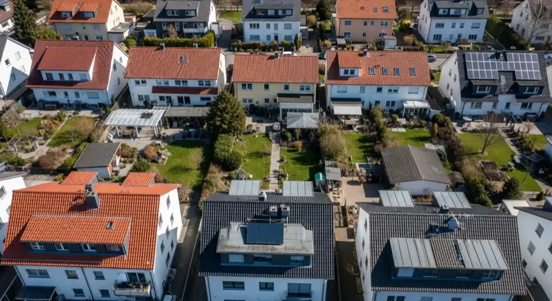 Aerial drone view of typical German residential neighborhood with mixed roof types, red and dark roof tiles, gardens visible, sunny day
